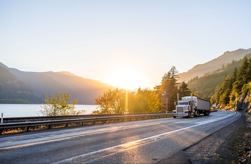 A white long-haul semi-truck is driving down a long highway alongside a river